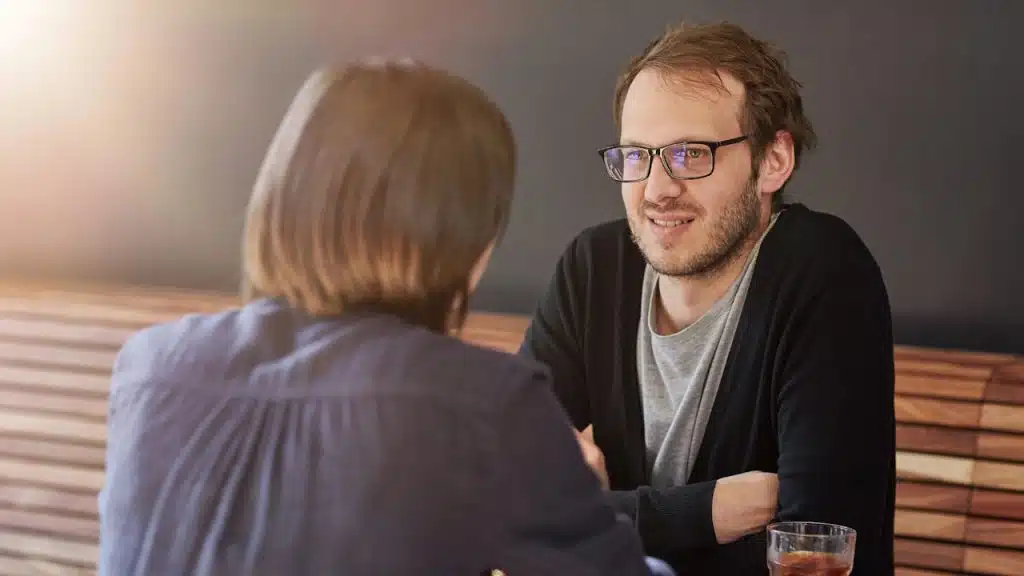 Man in glasses smiling across a table at another person.