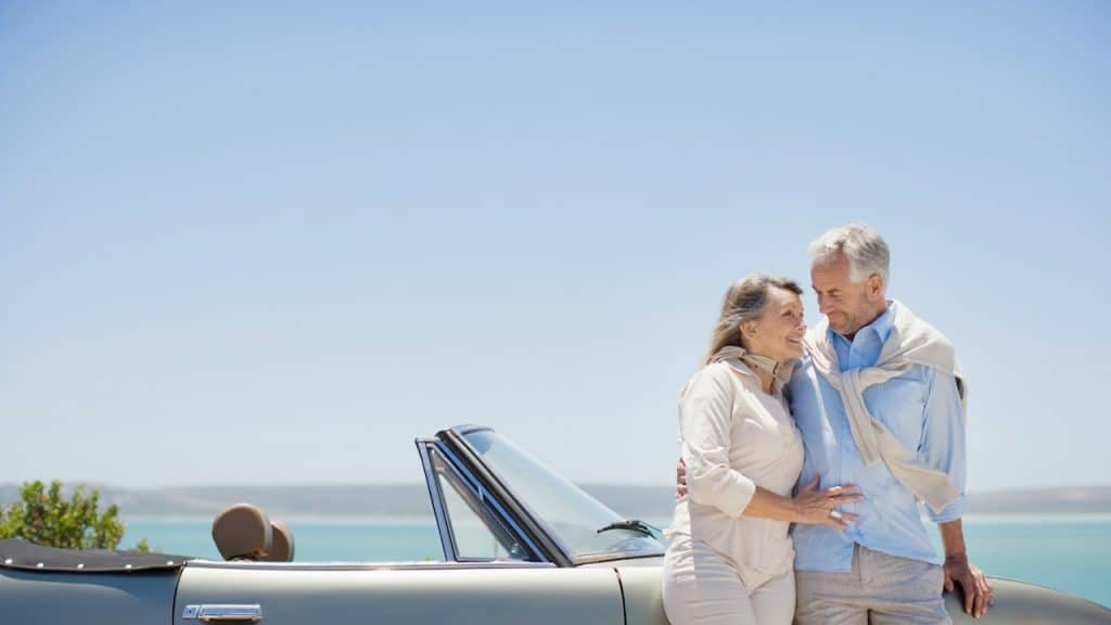 A man and a woman standing next to a luxury car beside a body of water.