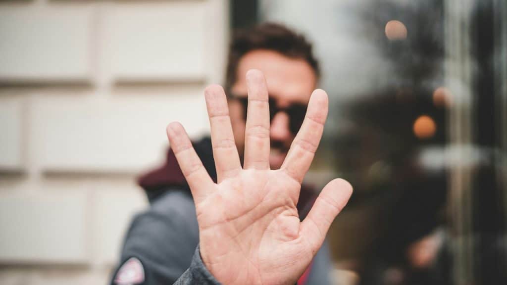 A man wearing sunglasses saying no by holding his hand up, palm outwards towards the camera.
