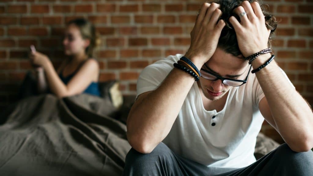 Distressed man sitting on a bed with his head in his hands, while a woman uses her phone in the background.