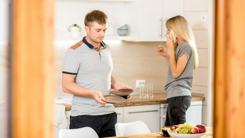 A man holds plates in a kitchen while a woman stands behind him talking on a phone.