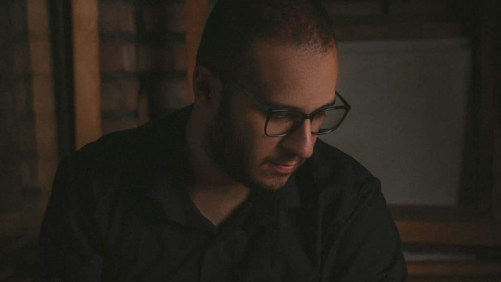 A man with short hair and glasses, wearing a dark shirt, looks down in a dim setting.