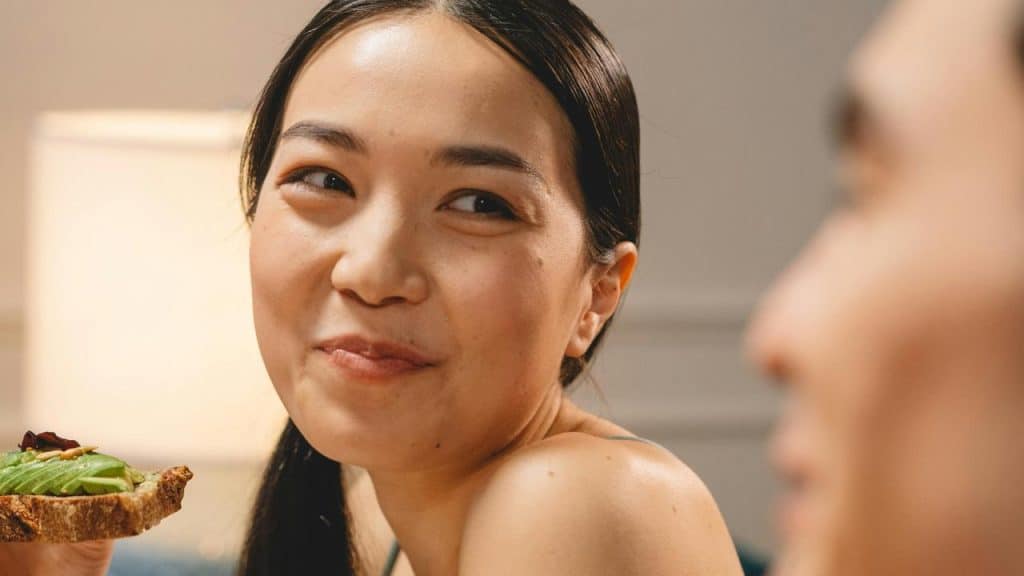 A woman smiling while holding a slice of avocado toast and looking at someone.