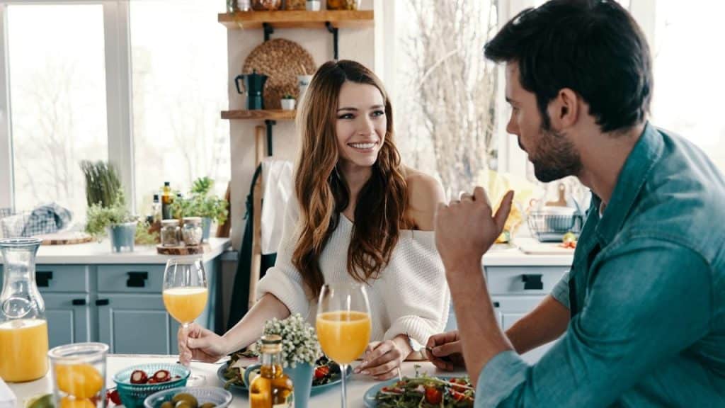 A smiling woman holds a glass of orange drink while talking to a man across a table.