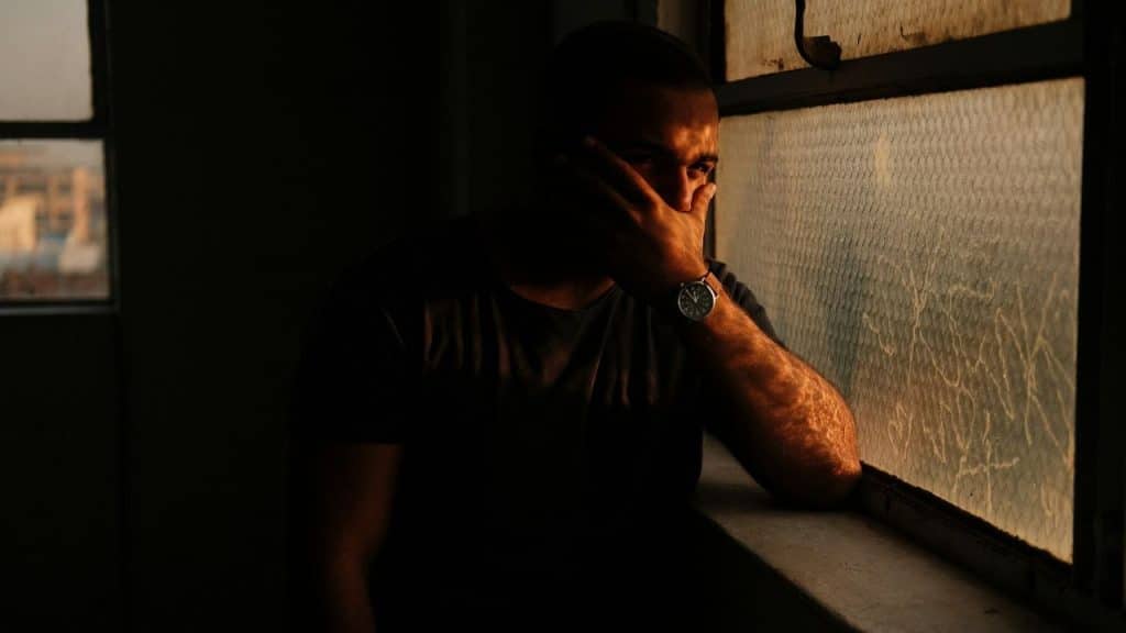A man in a dark shirt sits by a window, covering his face with one hand.