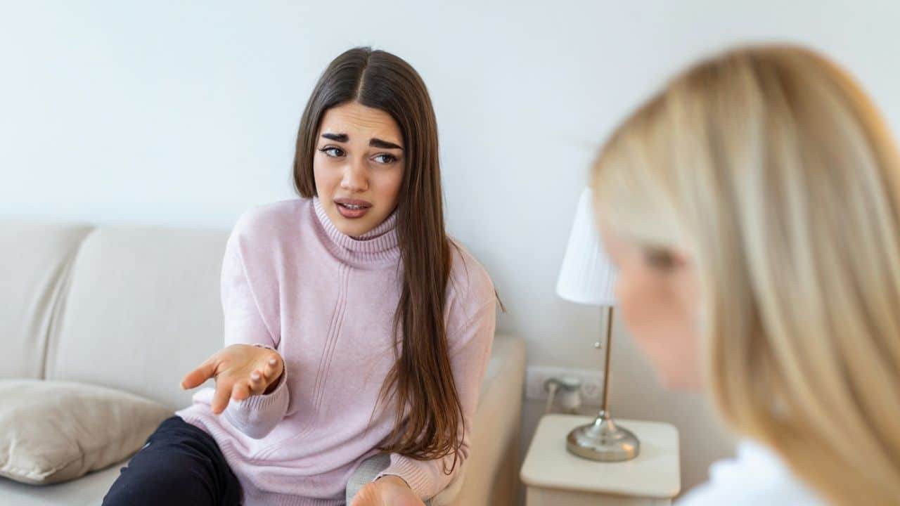 A woman in a pink sweater gestures with a worried expression while talking to a blurred person.
