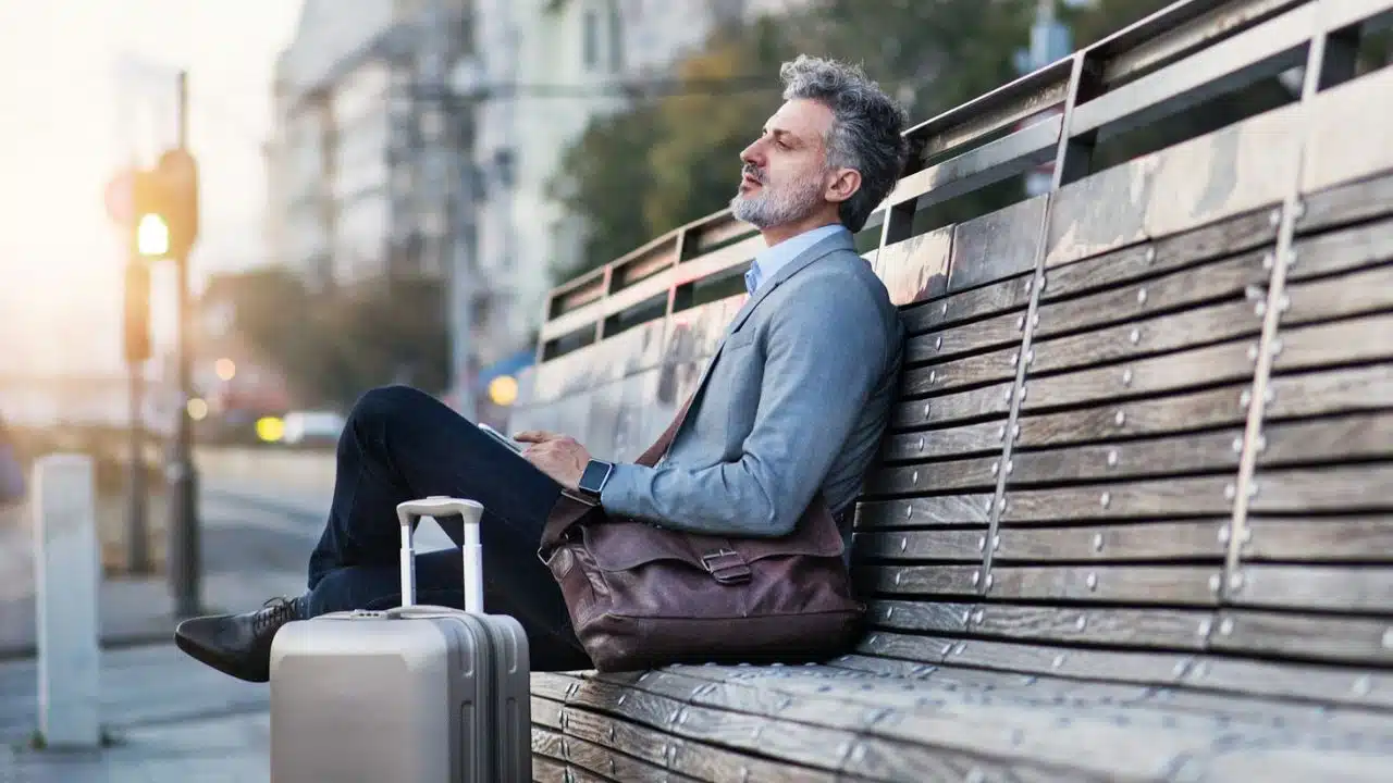 Bearded man relaxing on a wooden bench outdoors with a suitcase and briefcase nearby.
