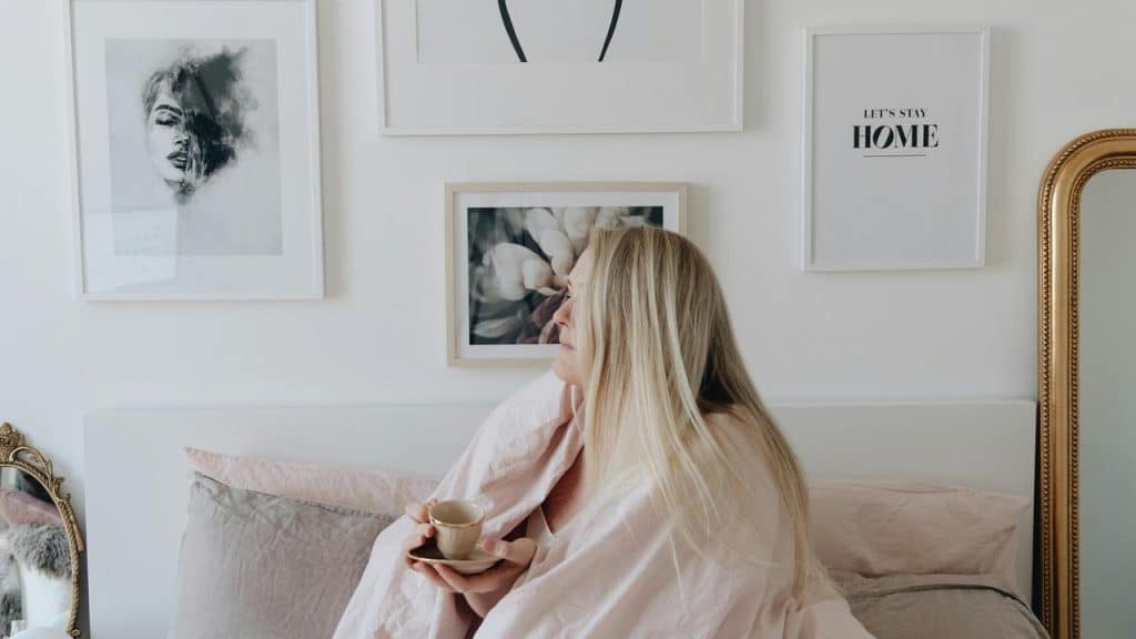A woman wrapped in a blanket sits in bed holding a cup and looking at wall art.