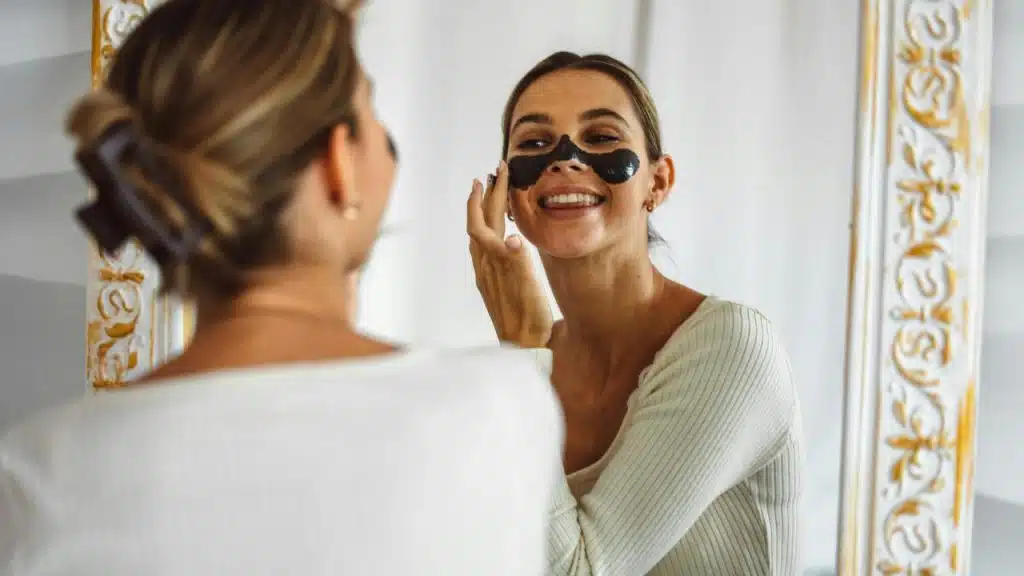 A woman smiling in the mirror while applying a black face mask.