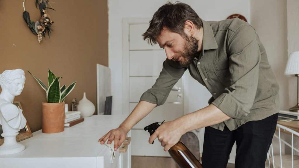 A man cleans a cabinet with a spray bottle and cloth in a home setting.