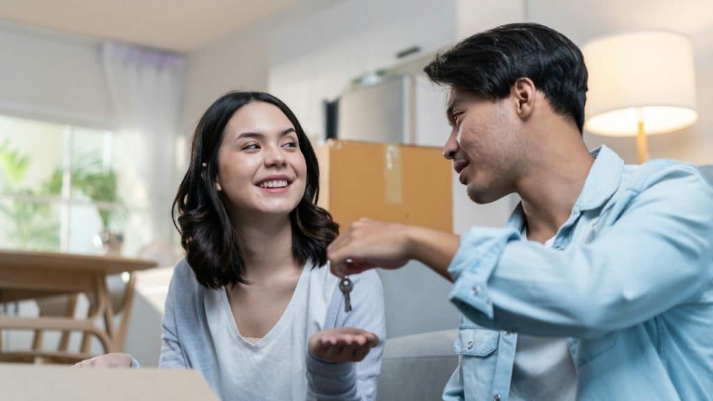 Man handing house keys to a smiling woman in a room surrounded by moving boxes.