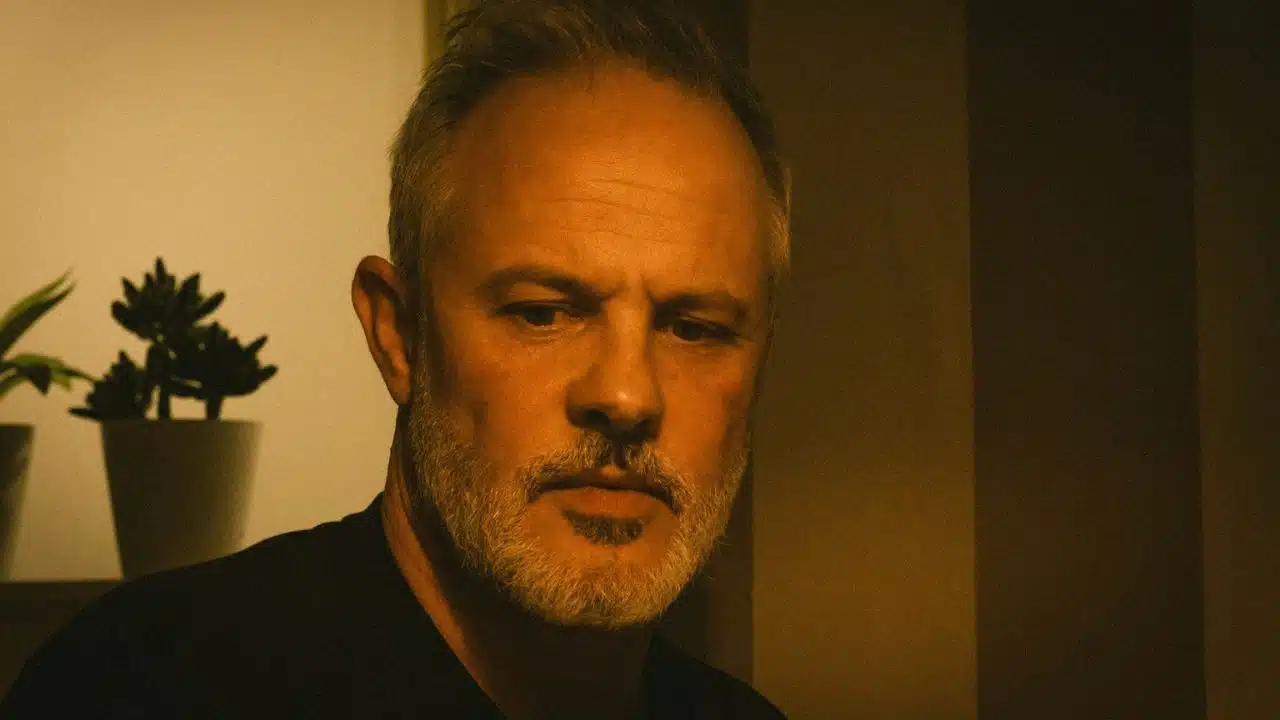 A serious, bearded man with gray hair is illuminated by warm, low light.