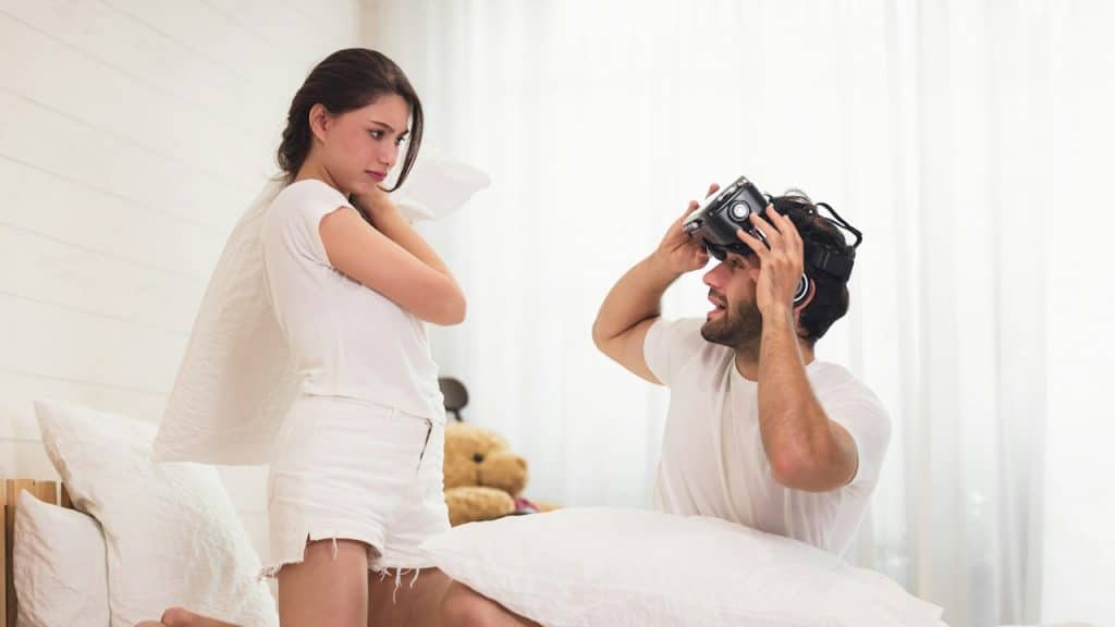 A woman with her husband on the bed, who is using his VR headset to play a video game.