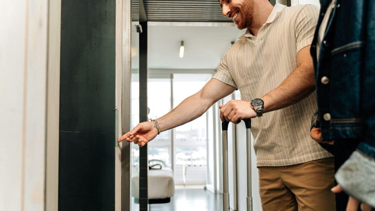 A smiling man pushes an elevator button while holding a luggage handle next to a person.