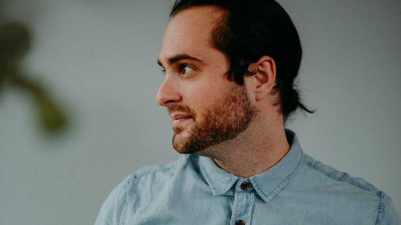 A man with a beard and long hair is looking over his shoulder against a light background.