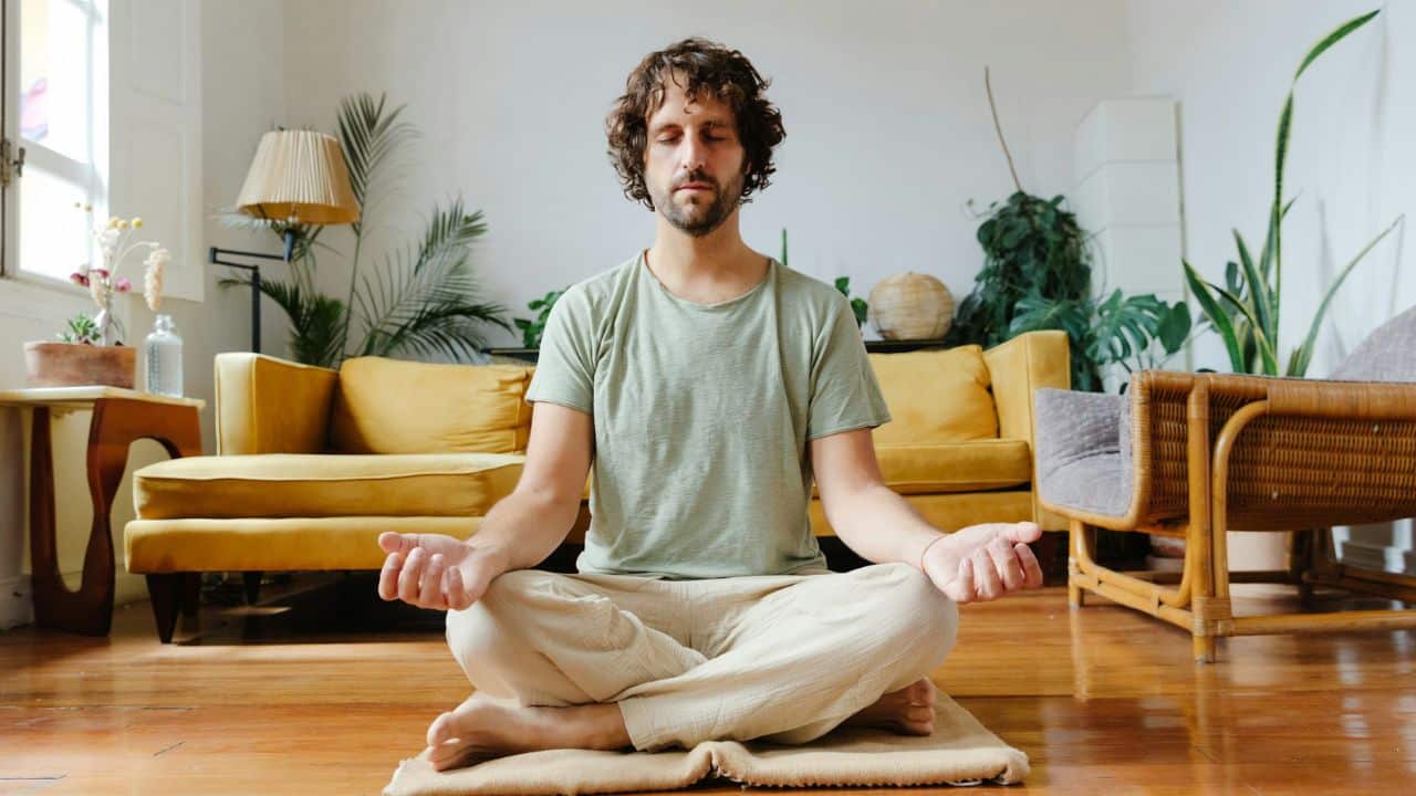 A man meditating cross-legged on a rug in a bright living room.