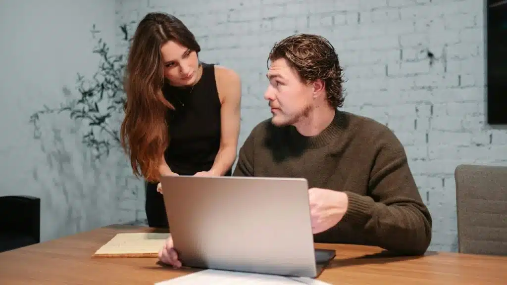 A woman leans over a man who is looking at a laptop on a wooden desk.