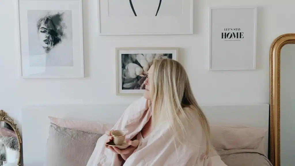 A woman wrapped in a blanket sits in bed holding a cup and looking at wall art.
