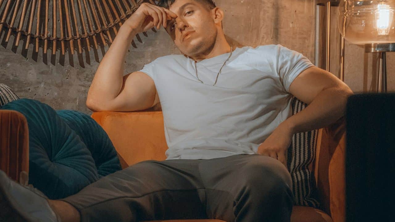 A muscular man in a white t-shirt and sweatpants sits on an orange chair.