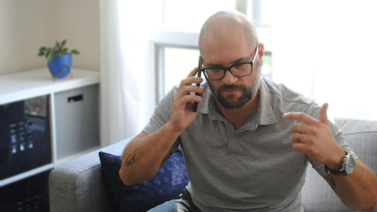 A bald man with glasses and a beard sits on a couch talking on a cell phone.