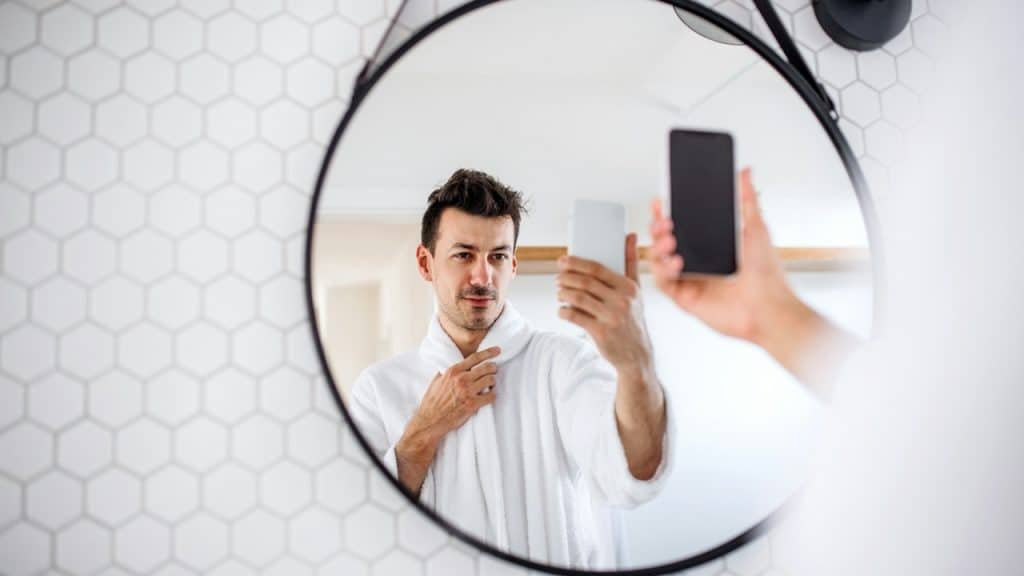 A man using his phone to take a selfie while looking at his reflection in a bathroom mirror.