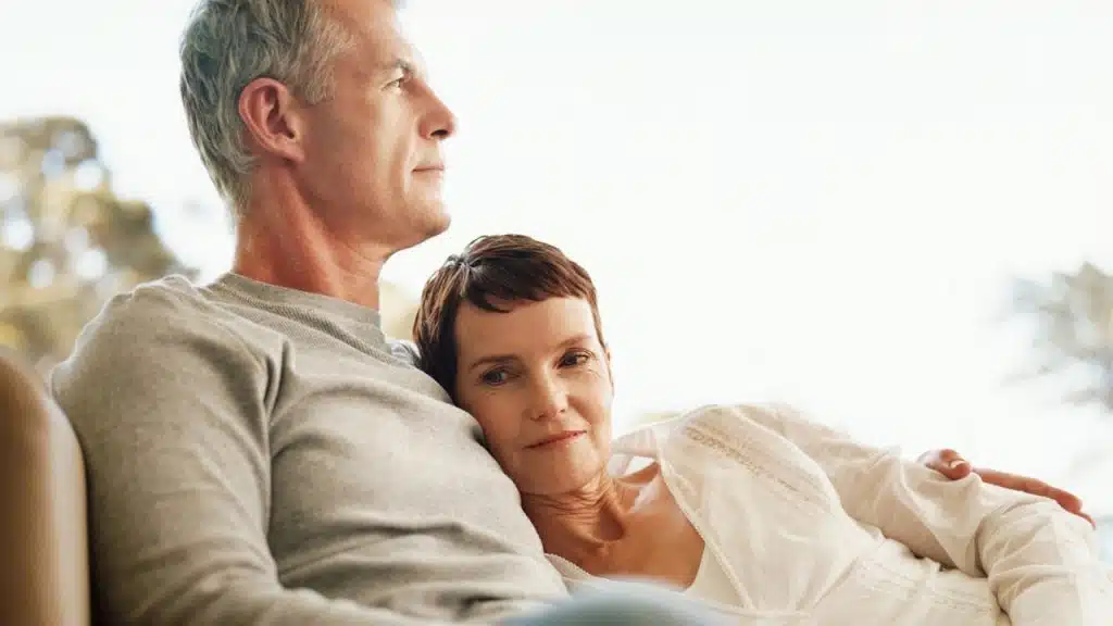 An older couple relaxes together indoors, with the woman resting her head on the man's shoulder.