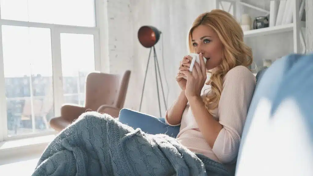 A woman sitting on a couch wrapped in a blanket, drinking from a mug.