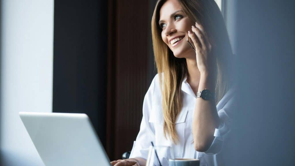 A smiling woman talking on a phone while working on a laptop.