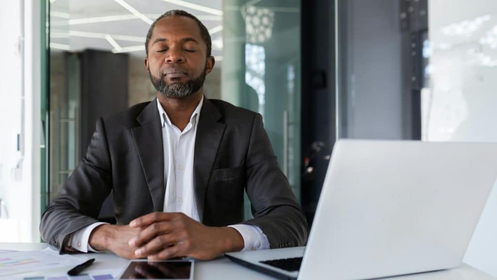 A man in a suit sitting at a desk with closed eyes, appearing to concentrate.
