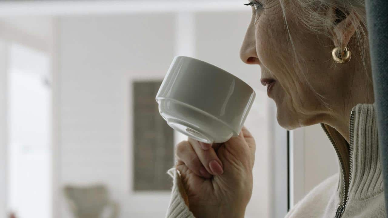 An older woman sipping from a white cup while looking out a window.