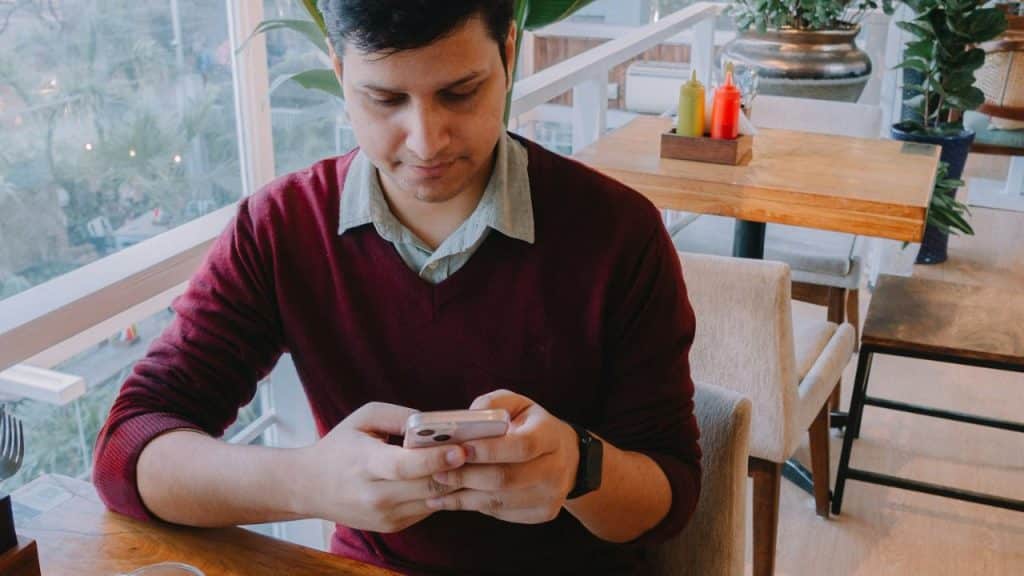 A man wearing a burgundy sweater looks intently at his cell phone at a restaurant table.