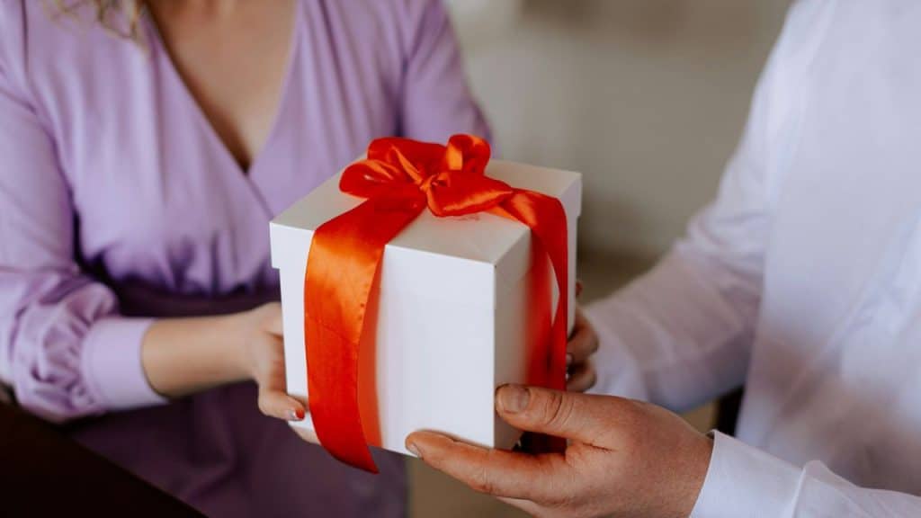 A white gift box tied with a red ribbon being passed from one person to another.