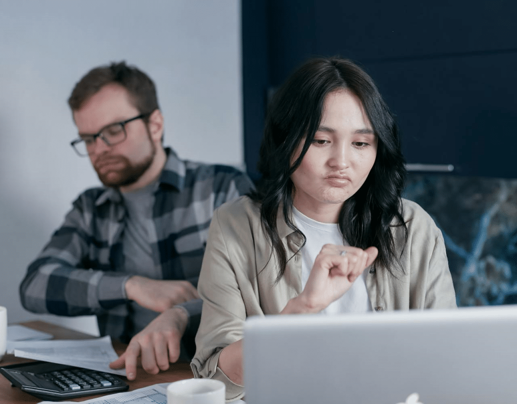 An angry woman is working on a laptop while a silent man is reading some documents.