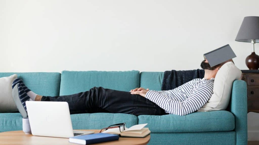 A man is lying on a turquoise sofa with a tablet covering his face; a laptop is nearby.