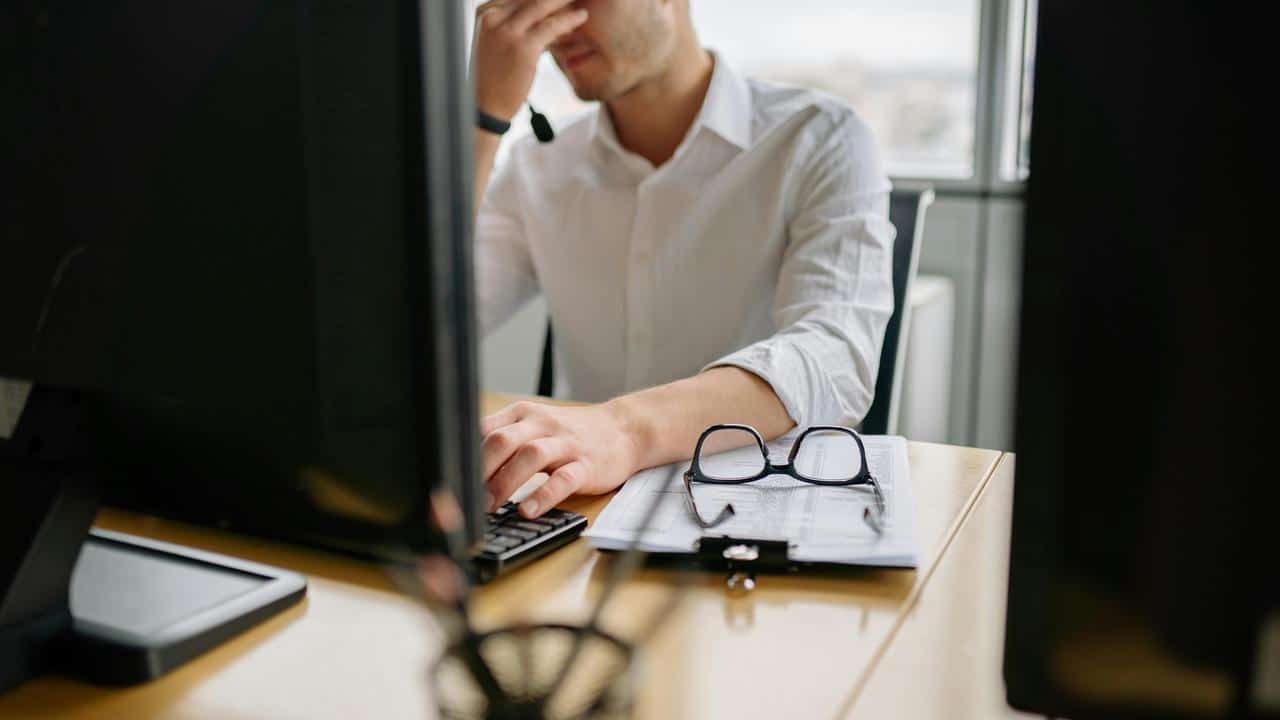 A man at a desk holding his face in frustration while working on a computer.