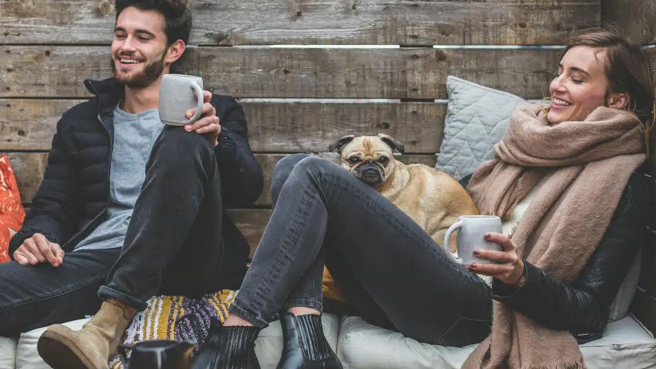 A couple relaxing with hot drinks and a small dog between them.