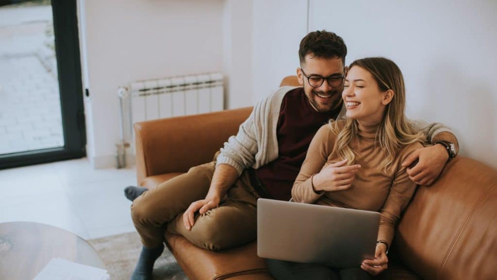 Smiling couple on a brown couch looking at a laptop together.