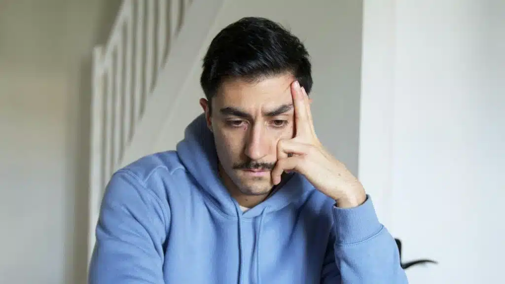 A man in a blue hoodie is sitting, leaning his head on his hand.