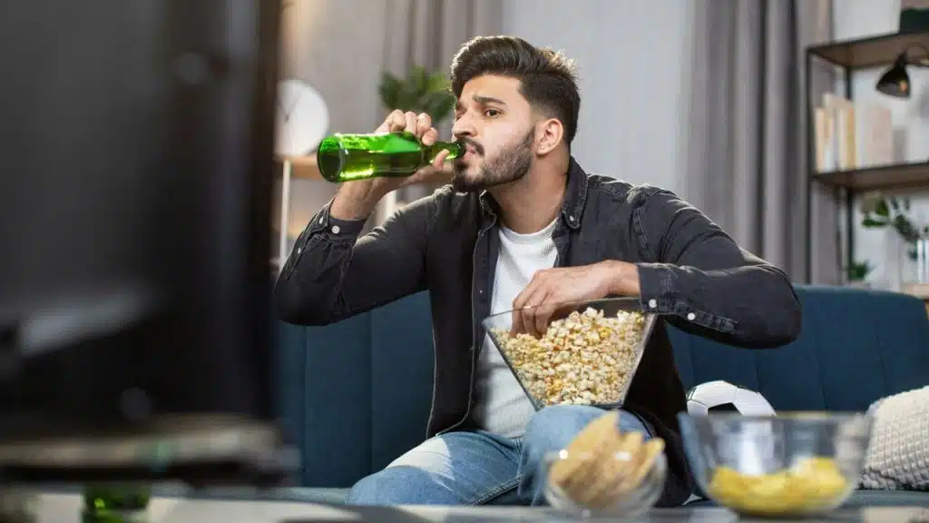 A man sits on a couch, drinking from a green bottle and eating popcorn.