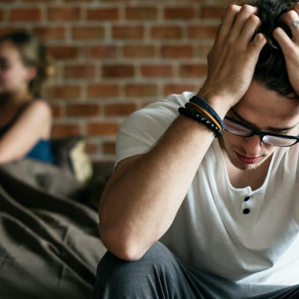 A distressed man is sitting on a bed with his head in his hands, while a woman uses her phone in the background.