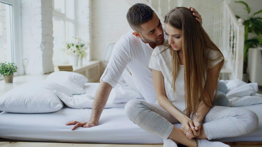 A man is comforting a sad woman in bed, holding her shoulder while she sits looking down.