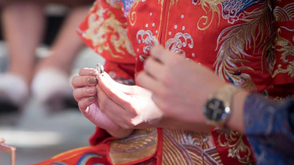 Close-up of hands during a wedding, with the person on the left holding an engagement ring while wearing traditional red and gold Chinese wedding attire.