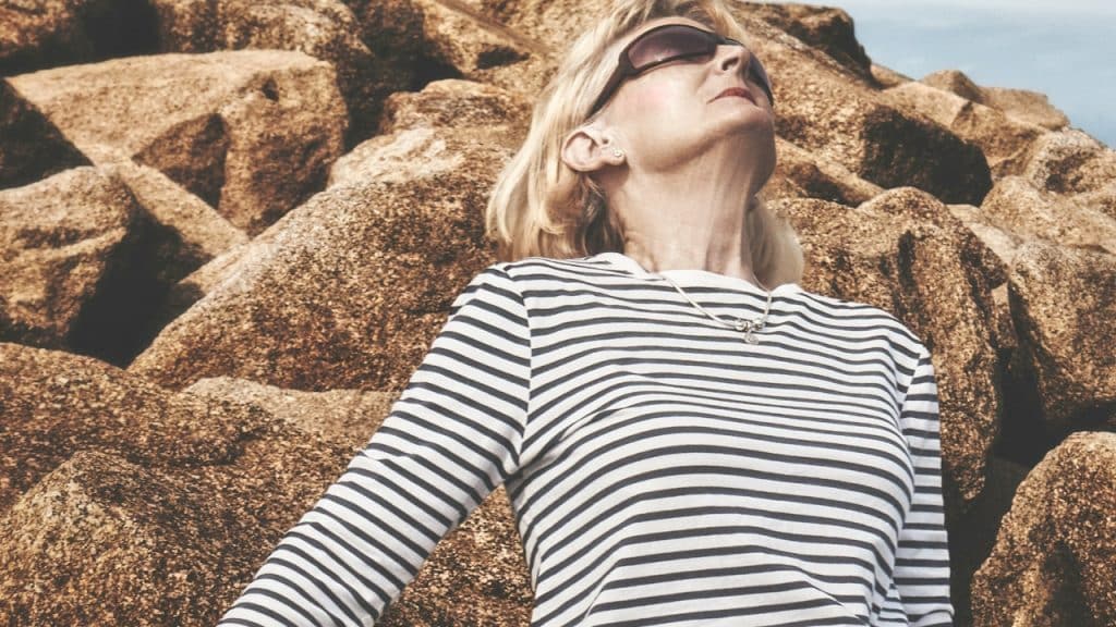 A woman wearing sunglasses is looking upwards while leaning against some rocks.