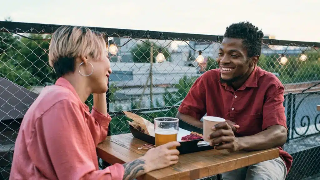 A happy couple sits at an outdoor table on a rooftop or balcony, smiling and talking while having drinks.