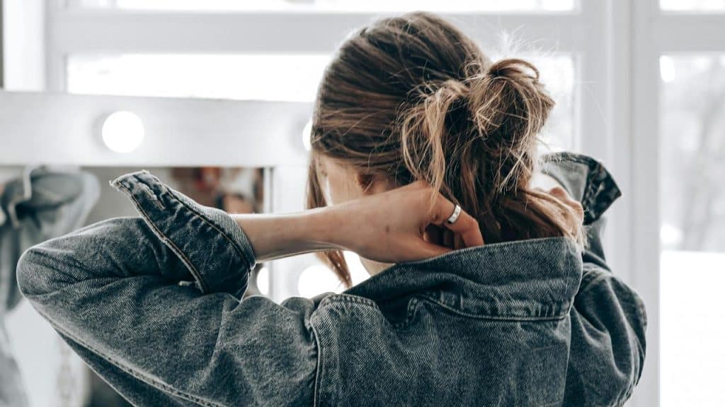 A woman in a denim jacket adjusts her hair while facing a mirror.