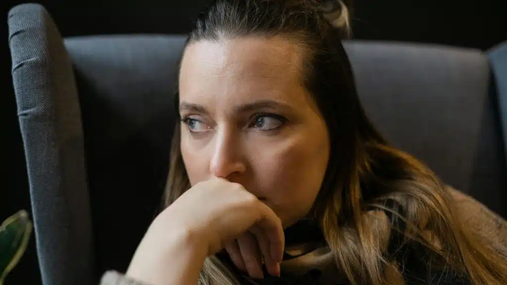 A woman sits with her hand near her face, looking thoughtful and concerned.