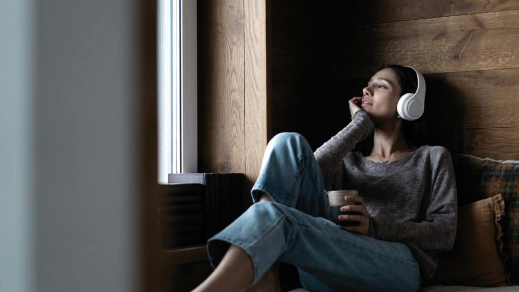 A woman relaxing by a window, listening to music with headphones and holding a cup.
