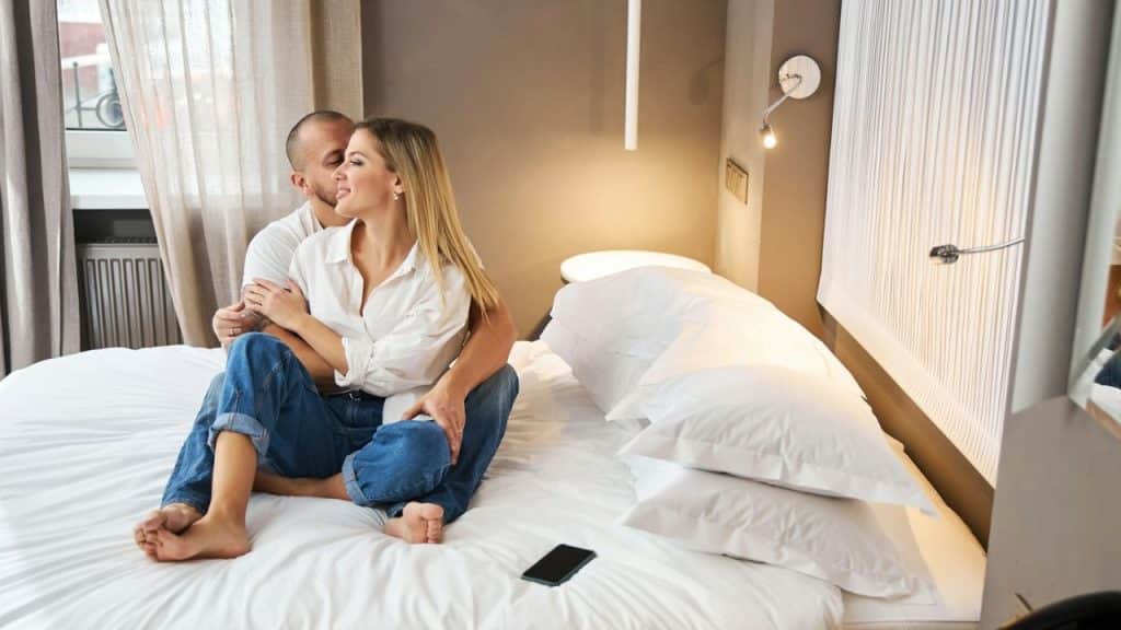 A man hugs a smiling woman from behind as they sit cross-legged on a bed indoors.