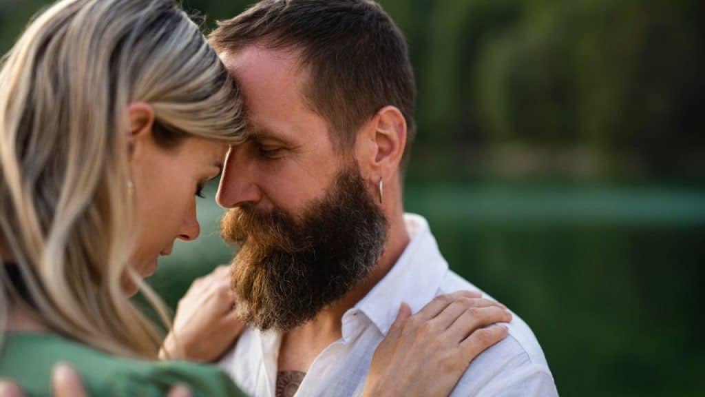A couple embracing outdoors, with the woman's forehead resting against the bearded man's forehead.