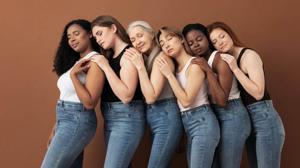 Six diverse women in jeans and tank tops leaning on each other with closed eyes.